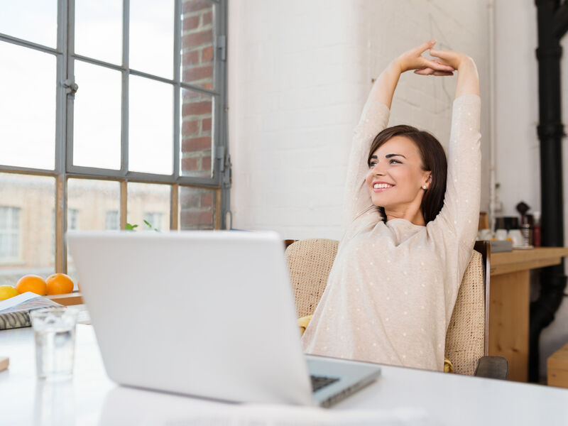 Happy relaxed young woman sitting in her kitchen with a laptop in front of her stretching her arms above her head and looking out of the window with a smile